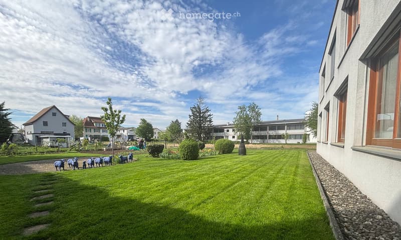 Green lawn, trees, shrubs, gravel path, sheep figurines, building in the background