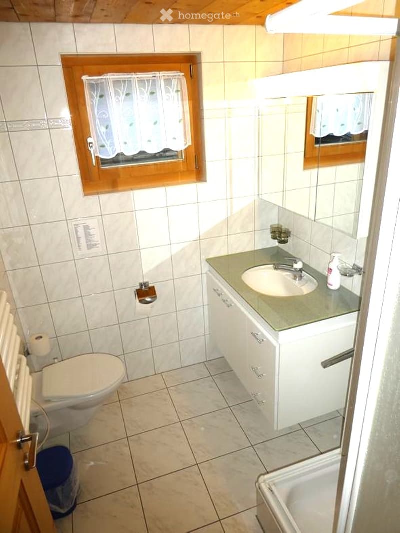 A small bathroom with white tiled walls and floor, featuring a toilet, a sink with a mirror above it, and a window with a white curtain. A soap dispenser and a small cabinet are also visible.