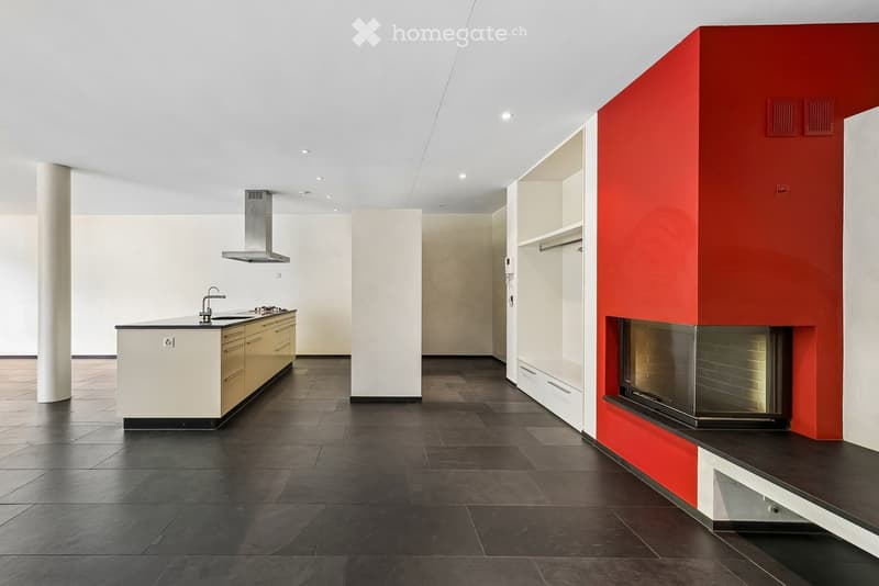 Modern kitchen with dark tiled flooring, white and beige cabinets, a central island with a sink and stove, and a built-in fireplace with a red accent wall.