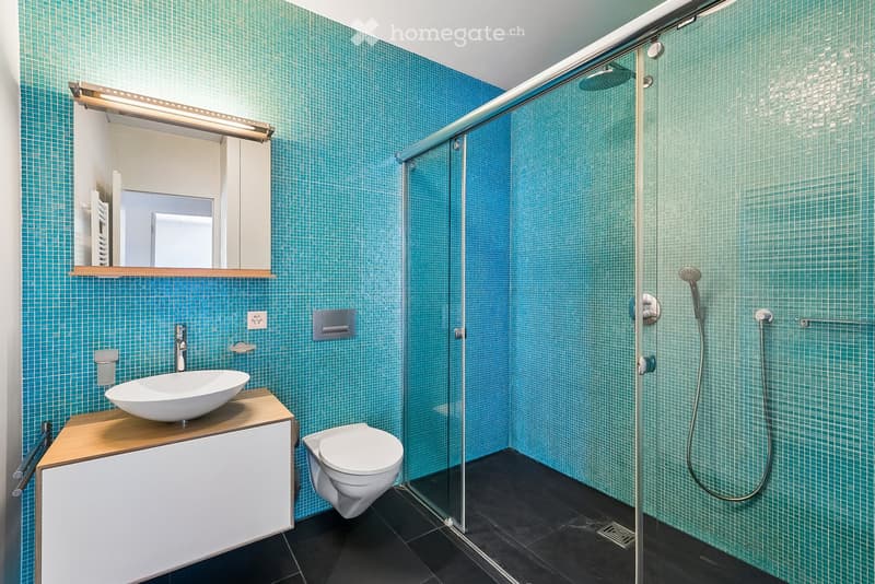 Modern bathroom with teal tiled walls, a glass-enclosed shower, a white sink on a wooden vanity, and a toilet.