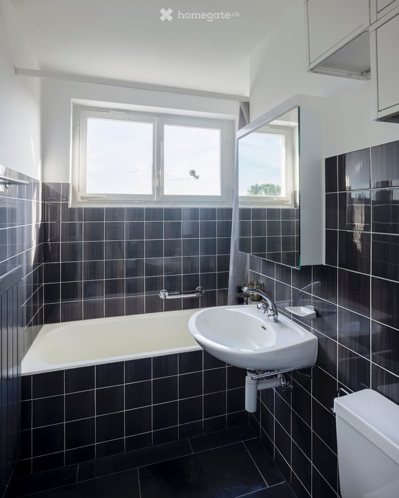 Modern bathroom with dark tiled walls and floor, featuring a bathtub, sink, mirror, and toilet. Large window provides natural light.