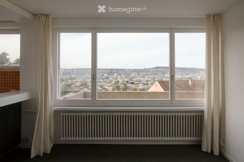 Empty room with large windows offering a cityscape view, radiator below the windows, and light-colored curtains on either side.
