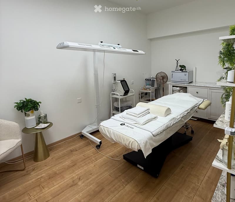 A well-organized massage or treatment room with a massage table, a small round table, a chair, a fan, a towel warmer, a screen, and plants.