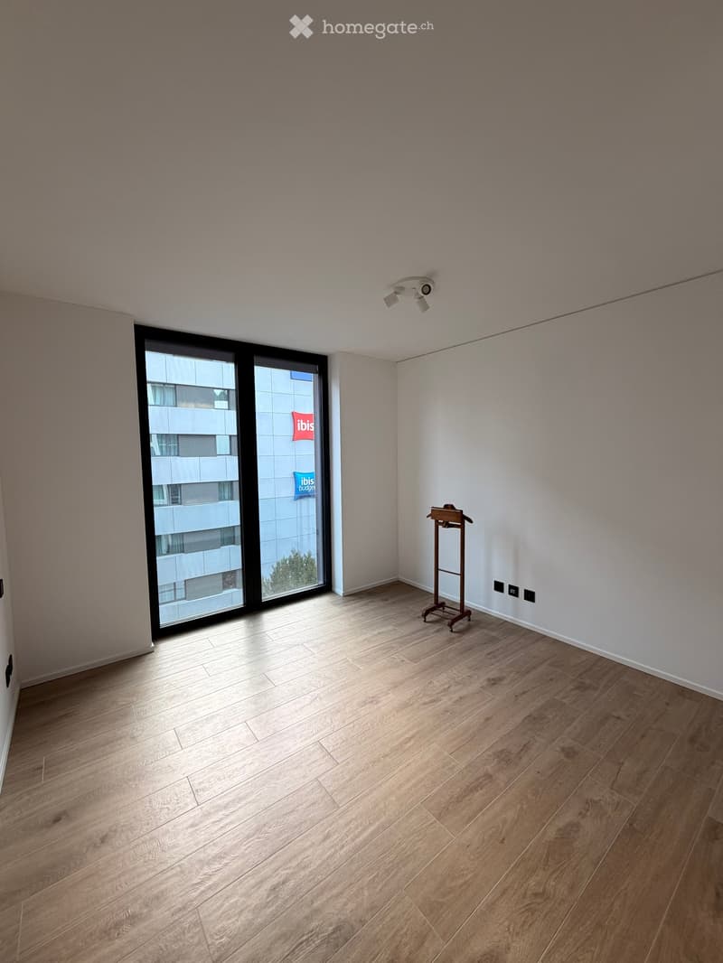 Empty room with light wooden flooring, white walls, and a large sliding glass door leading to a balcony. A small wooden coat rack is positioned near the wall.