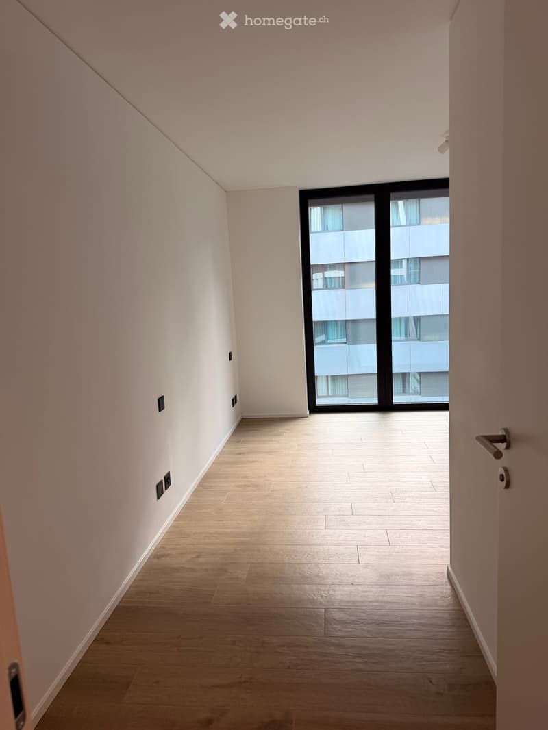 Empty hallway with light wooden flooring, white walls, and a large sliding glass door leading to a view of another building.