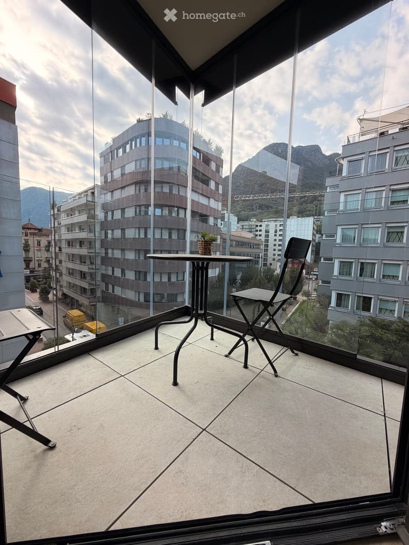 Balcony with tiled floor, glass railing, small table, chairs, and potted plant, overlooking cityscape and mountains.