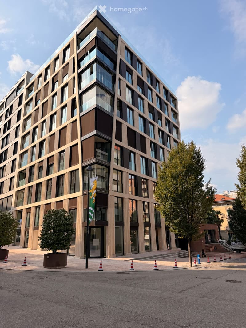 Modern multi-story building with large windows and a glass facade, surrounded by trees and traffic cones.