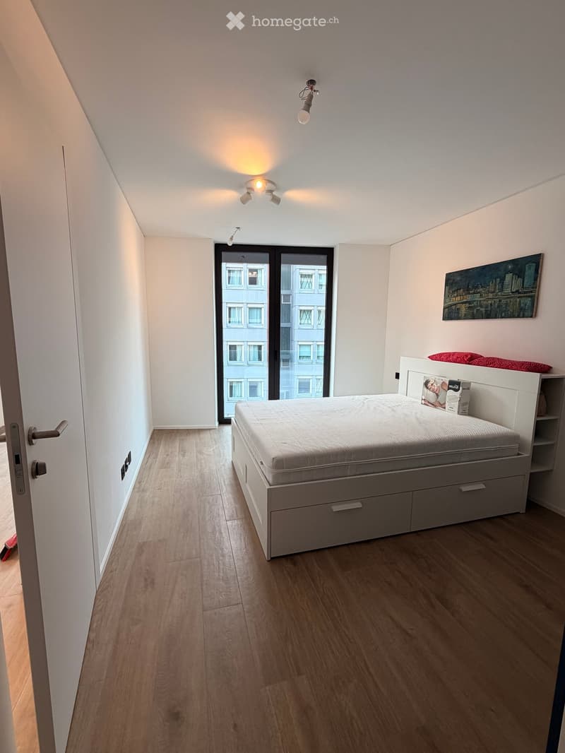 Modern bedroom with a bed, nightstand, and artwork on the wall. The room features wooden flooring and large windows with a city view.