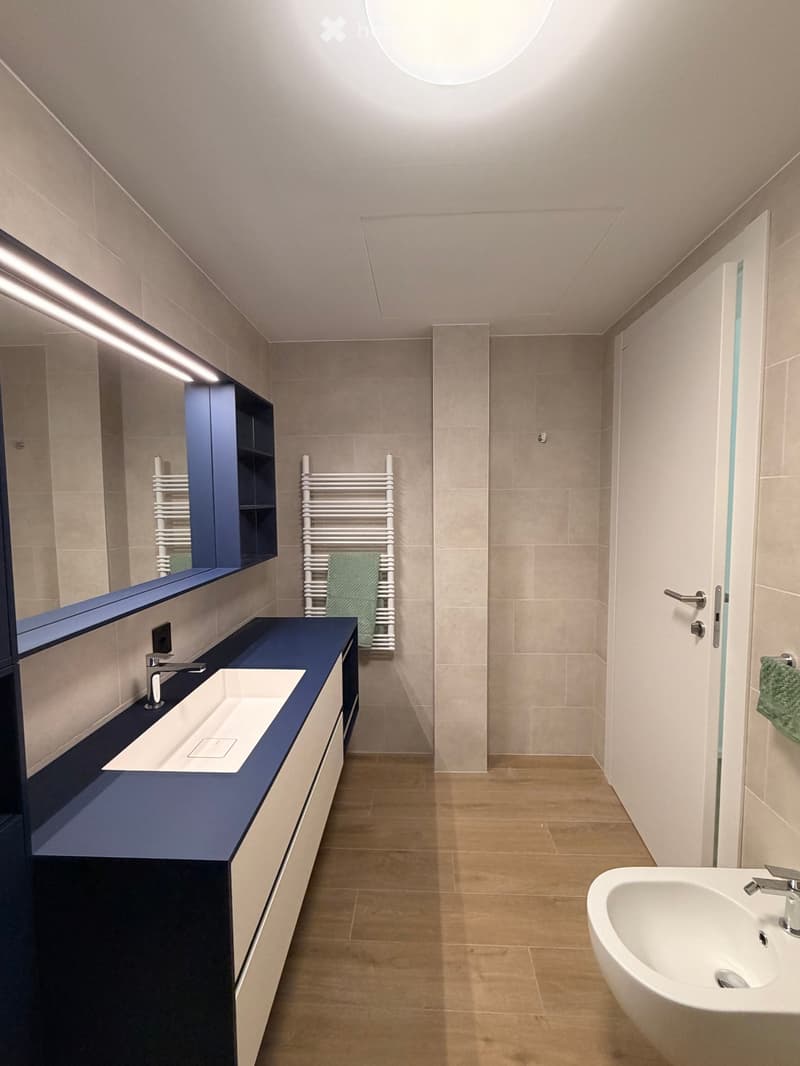 Modern bathroom with beige tiles, dark blue vanity, white sink, and a large mirror with built-in lighting. A towel rack with a green towel is visible.