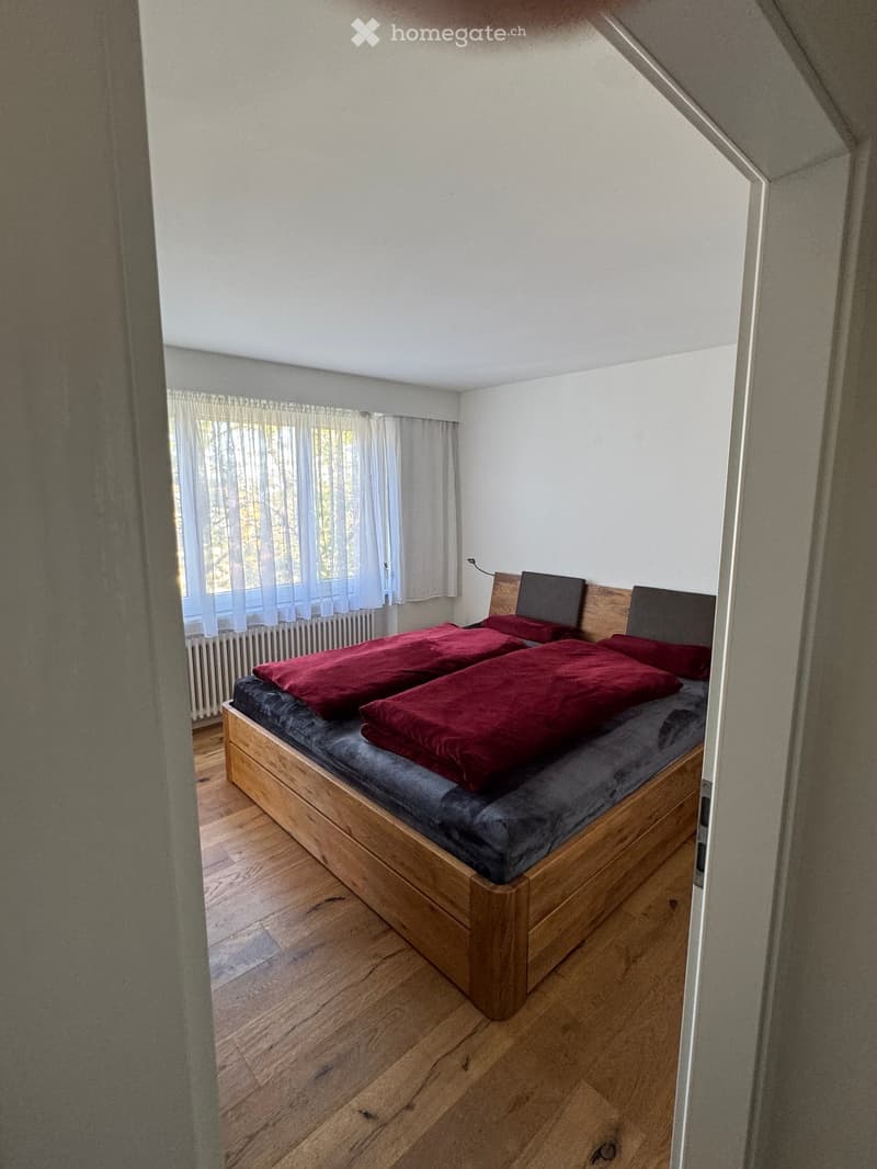 A small bedroom with a wooden bed, red pillows, and a window with sheer curtains. The room has wooden flooring and white walls.