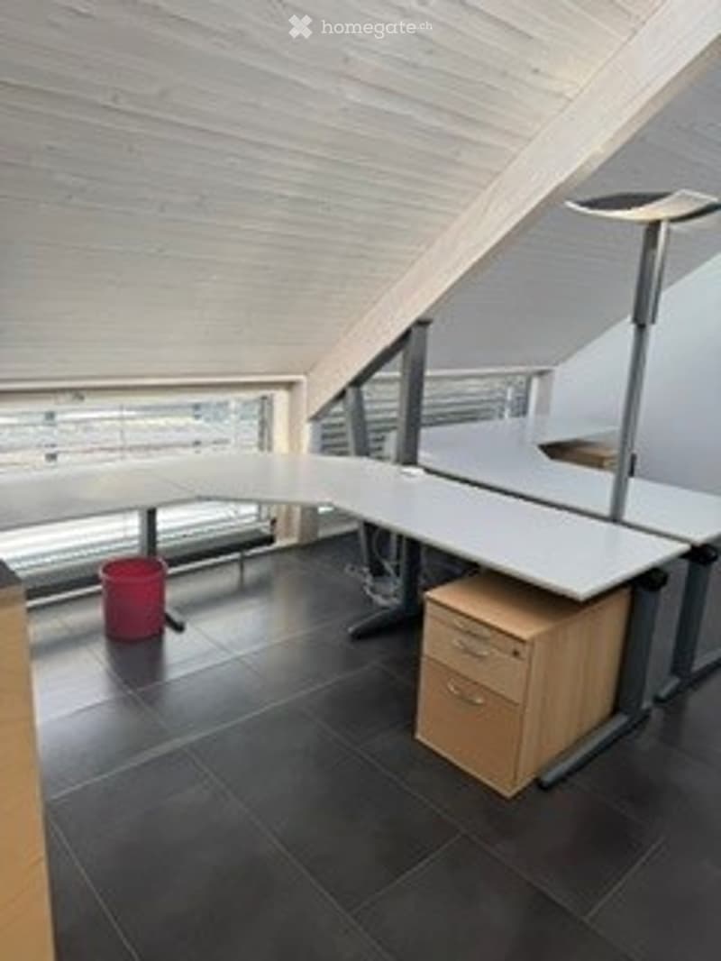 Modern, minimalist office space with a sleek white desk, ergonomic chair, wooden cabinet, and a small red trash bin. The room features exposed wooden beams on the ceiling and large windows allowing natural light to fill the space.