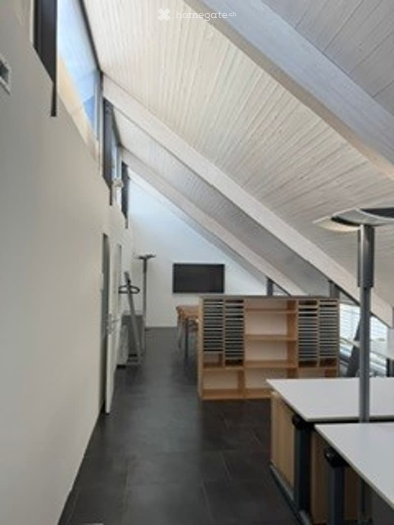 Modern, open-plan attic space with exposed wooden beams, dark flooring, and built-in shelving units. Includes a small table, chairs, and a screen.