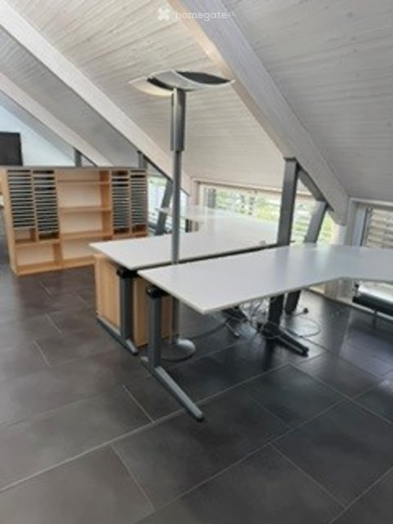 Modern office space with a large, curved white desk, ergonomic chair, and wooden bookshelf. The room features exposed wooden beams on the ceiling and dark tiled flooring.