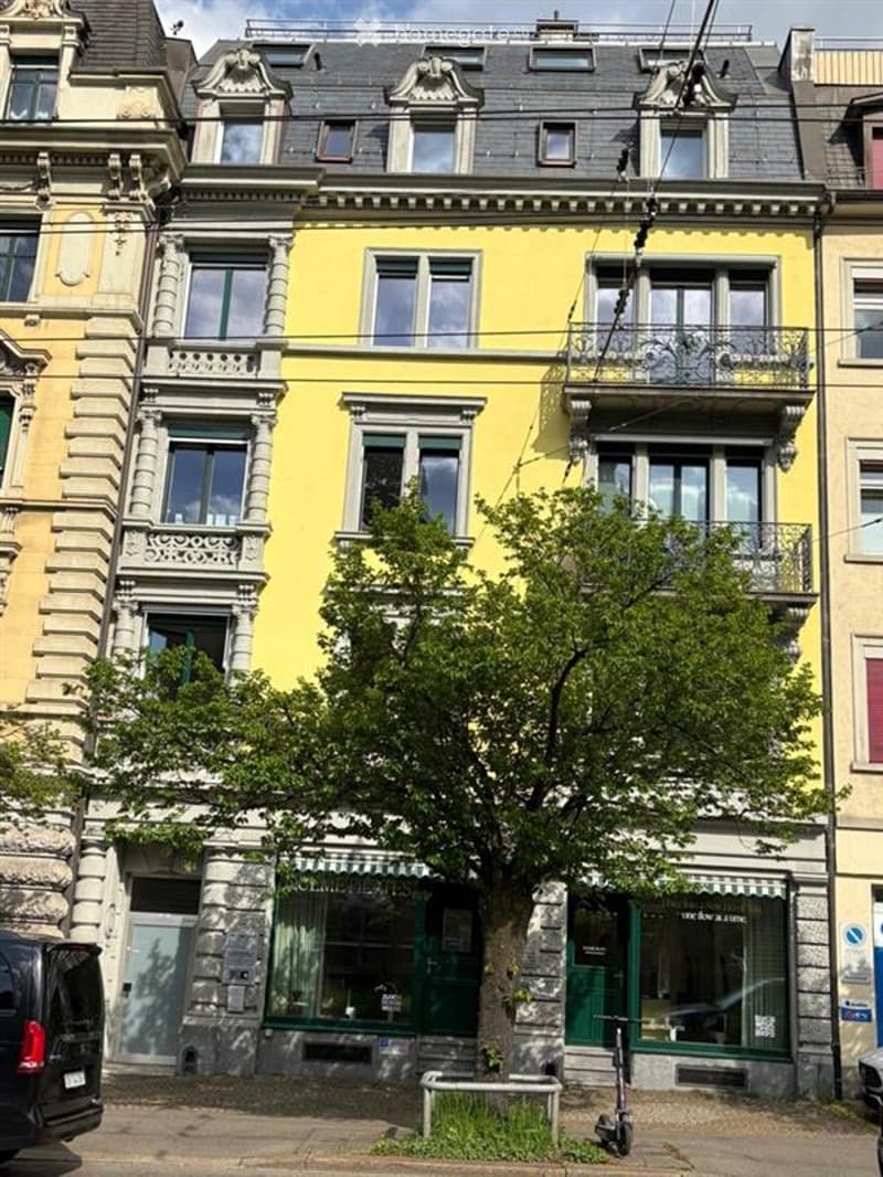 A multi-story residential building with yellow and grey facades, ornate balconies, and large windows. The ground floor features commercial storefronts with green doors and glass windows.
