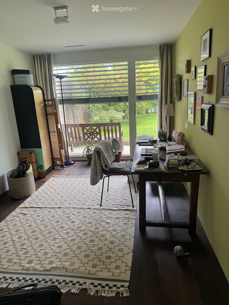 A cozy, well-lit room with a wooden floor, a patterned rug, a desk with a lamp, a chair with a blanket draped over it, and a bench visible through the window. The room features several picture frames on the walls and a small plant on the floor.