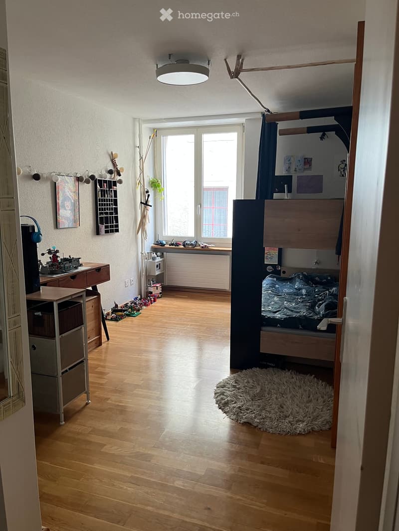 A well-lit bedroom with wooden flooring, a lofted bed, a desk with a chair, a storage unit, a large window with a curtain, and a shaggy rug on the floor.