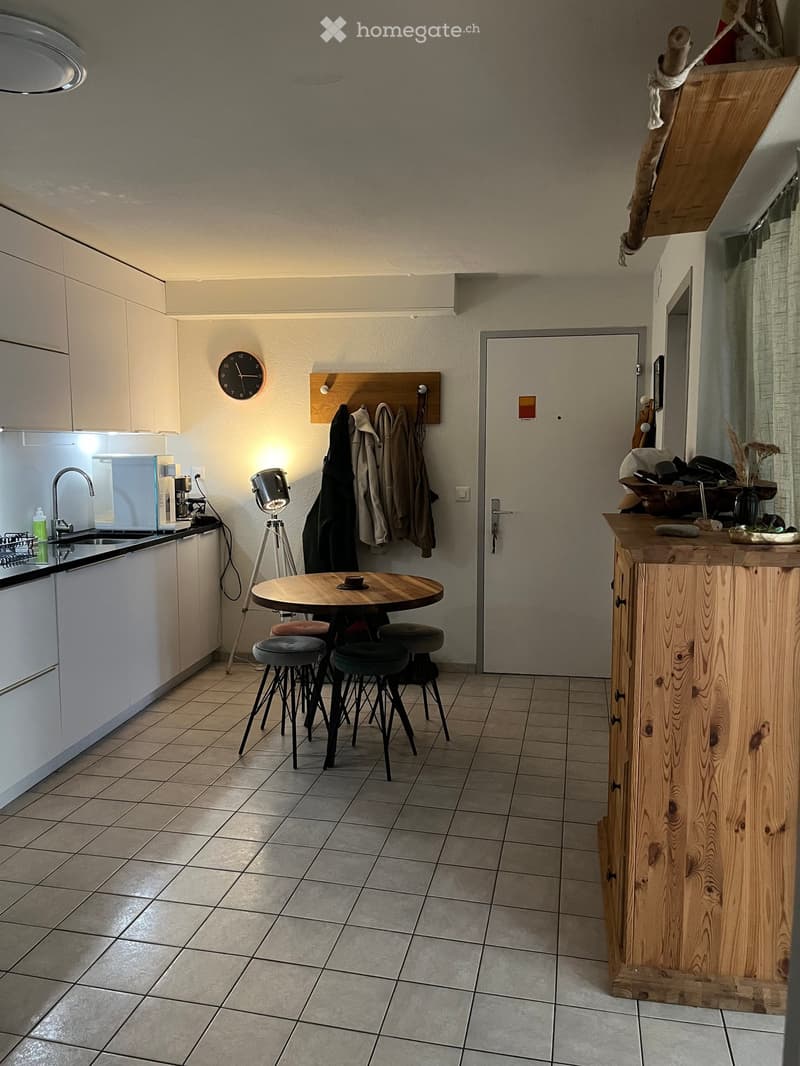 Modern kitchen with white cabinets, a wooden island, a round table with four stools, a sink with a faucet, and a coat rack with jackets.