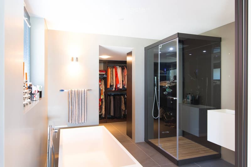 Modern bathroom with a large bathtub, glass-enclosed shower, and a walk-in closet with hanging clothes. The space features a towel on a rack, a sleek faucet, and a small shelf with toiletries.