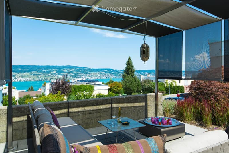A modern outdoor terrace with a panoramic view of a city and water, featuring a sofa, a small table with wine and glasses, and decorative cups.