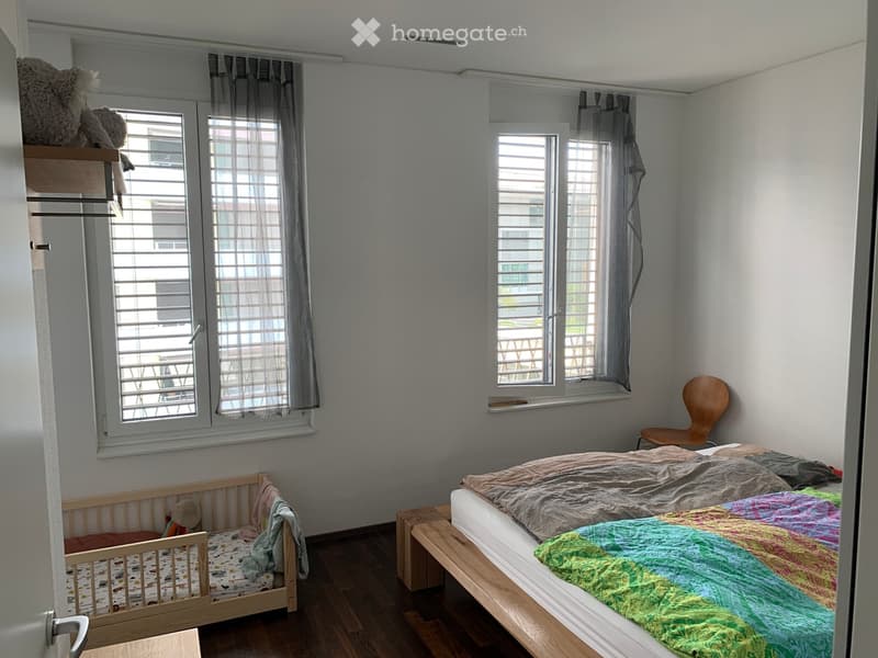 A small, cozy bedroom with a double bed, a wooden toy bed, a wooden chair, a shelf with toys, and large windows with blinds and curtains.