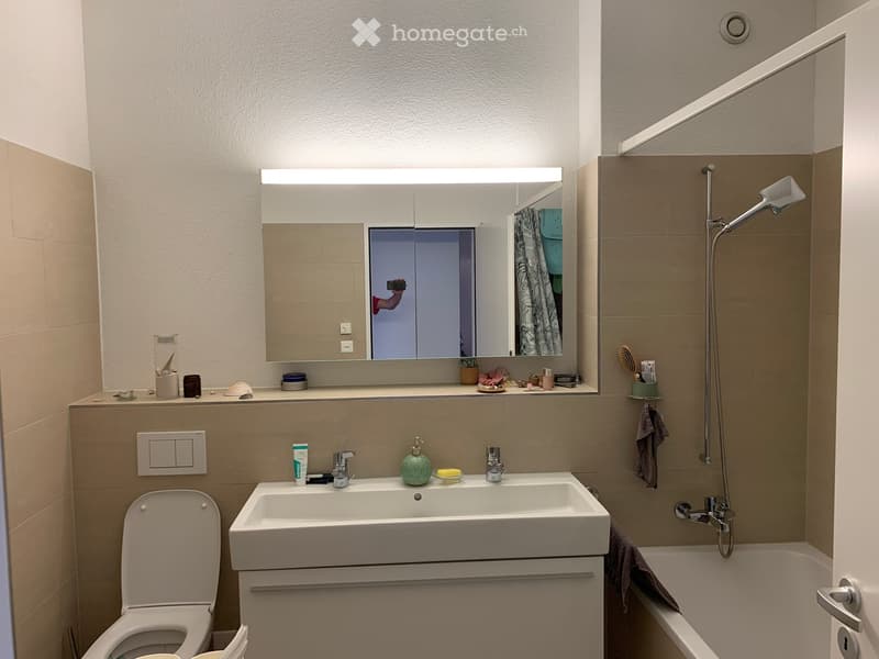 Modern bathroom with a white sink, toilet, bathtub, and a large mirror with built-in lighting. The space features beige tiles and a sleek, contemporary design.