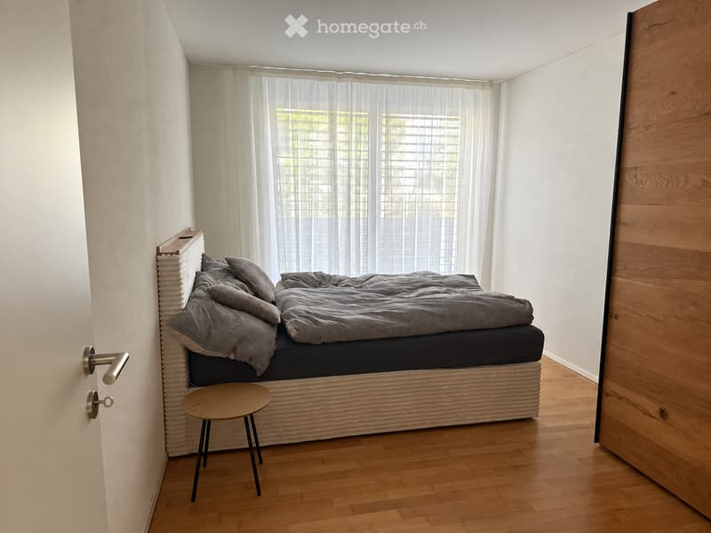 Modern bedroom with a wooden bed frame, grey bedding, a small wooden stool, and large windows with sheer curtains.