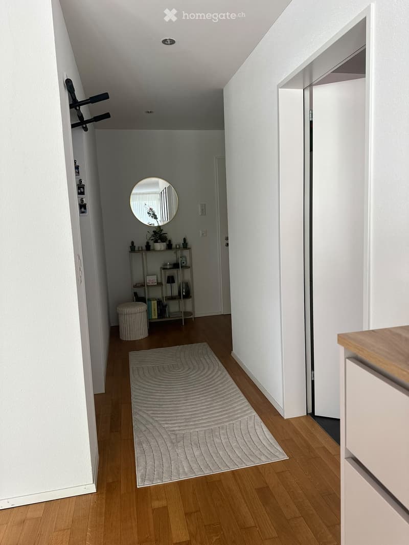 A narrow hallway with wooden flooring, white walls, and a grey patterned rug. A small shelf with books and decorative items is visible, along with a round mirror on the wall.