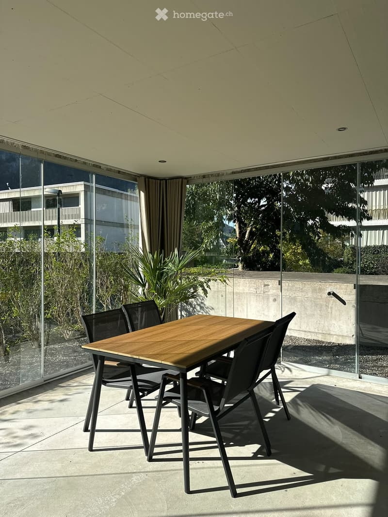 Modern outdoor dining area with a wooden table and four black chairs, large glass walls, and a view of greenery and buildings.