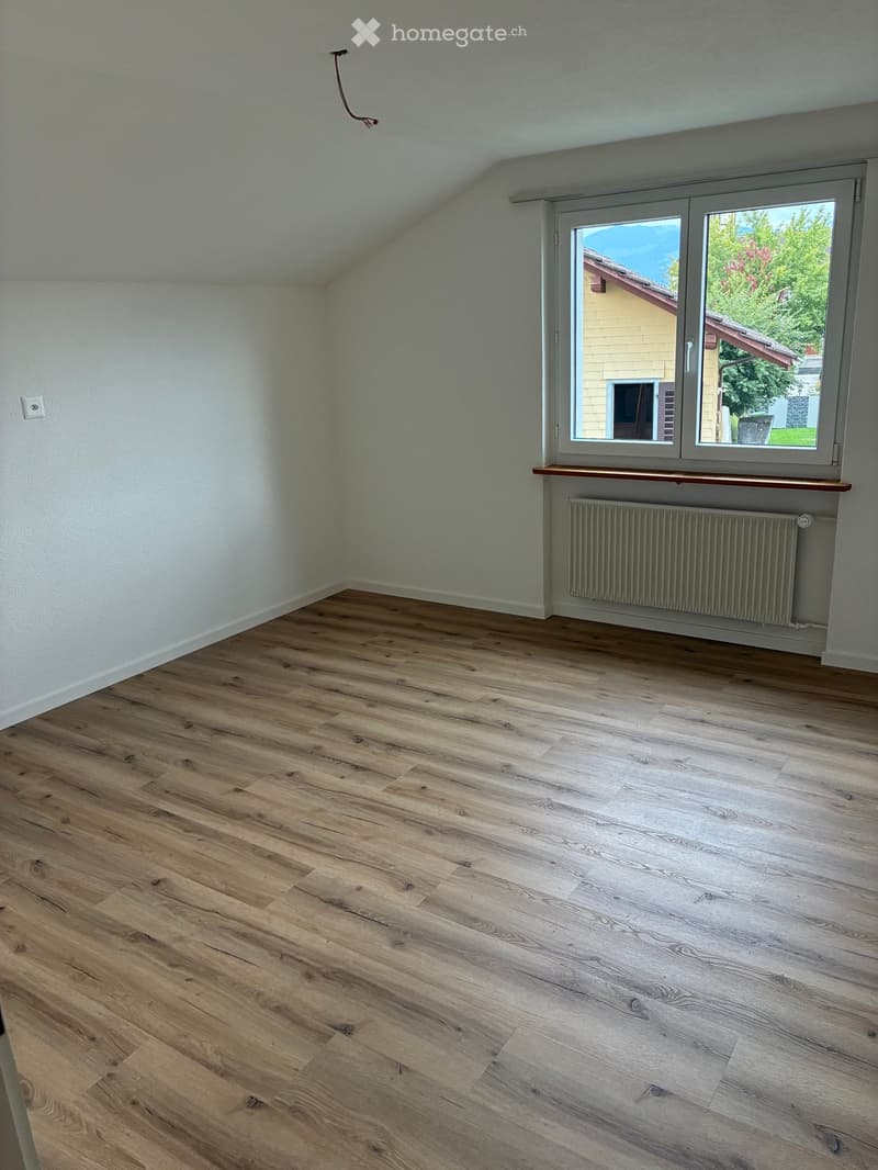Empty room with wooden flooring, white walls, large window with a view of a neighboring house, radiator, and a small wooden shelf under the window.