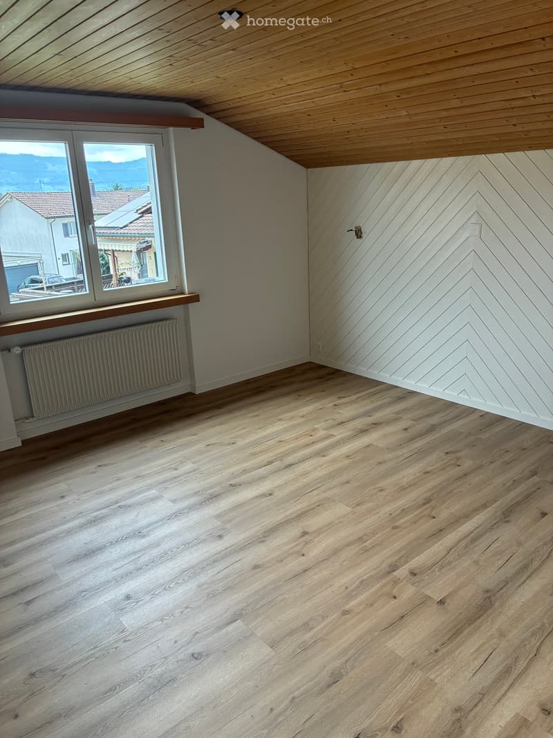 Empty room with wooden flooring, white walls, large window with view, radiator, and wooden ceiling beams.