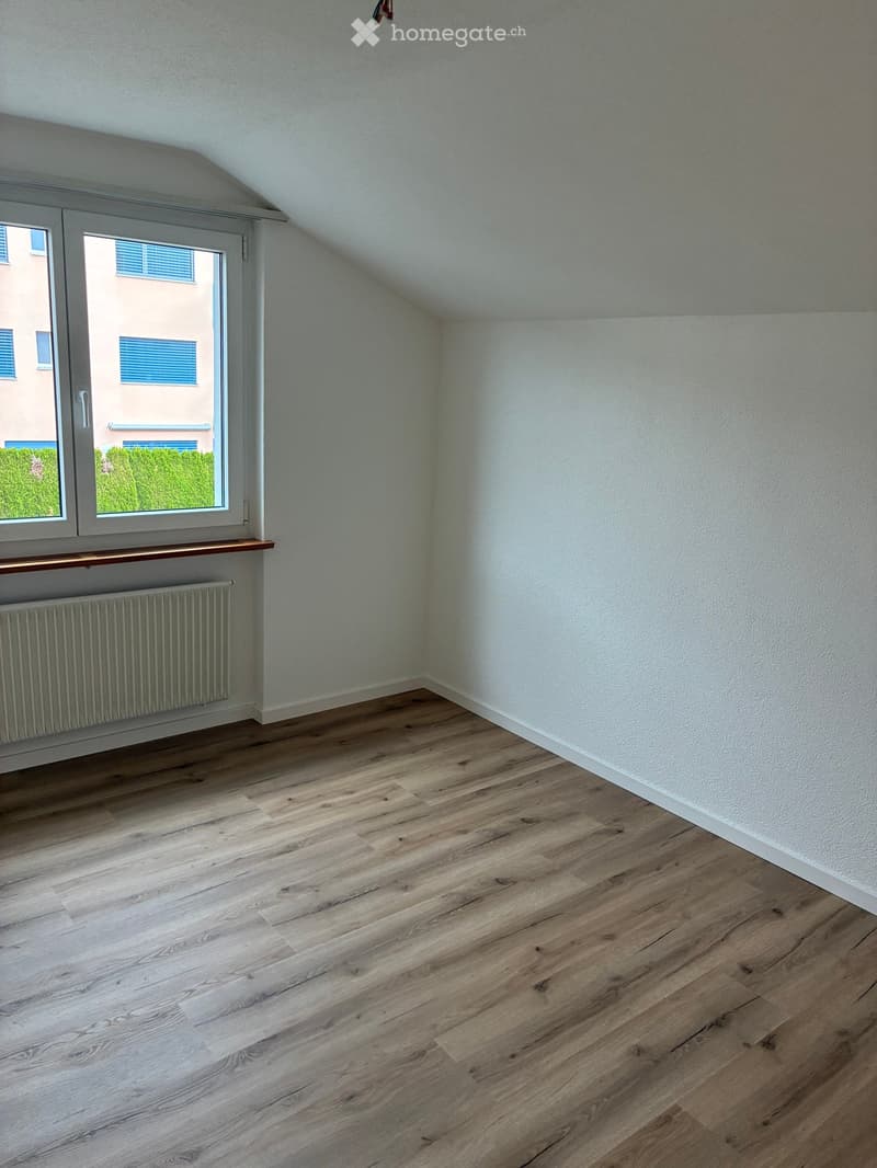 Empty room with wooden flooring, white walls, and a large window with a view of a neighboring building and greenery.