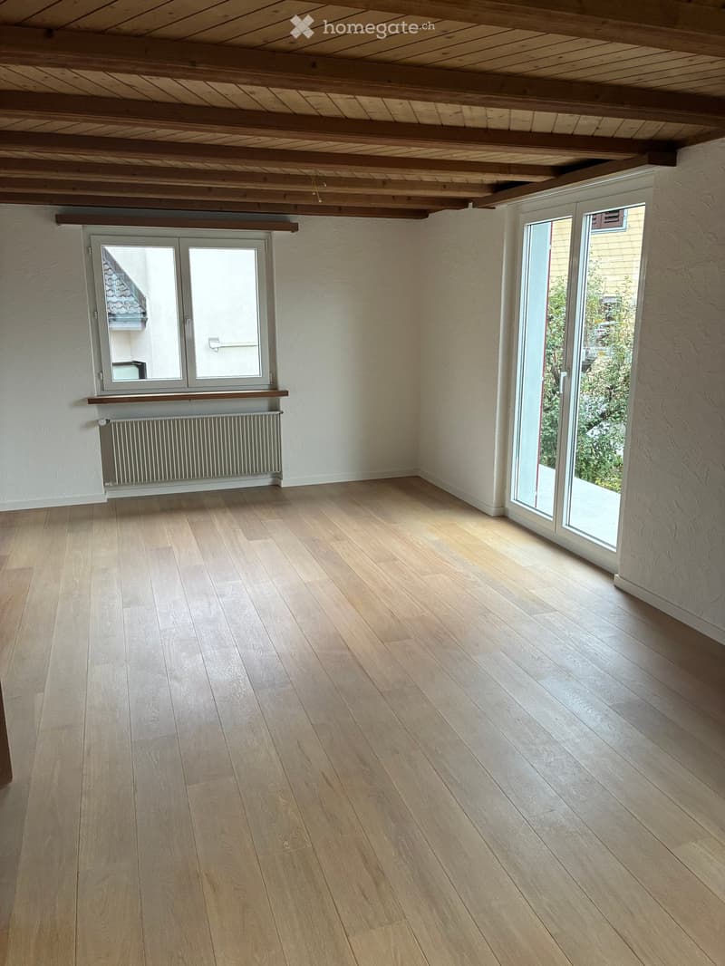 Empty room with wooden flooring, white walls, exposed wooden ceiling beams, and large windows with glass doors leading outside.