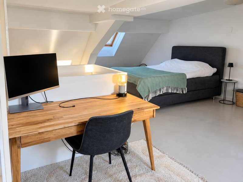 Modern bedroom with a large bed, desk with a screen, chair, and a small side table with a lamp. The room features a skylight window and a cozy green throw on the bed.