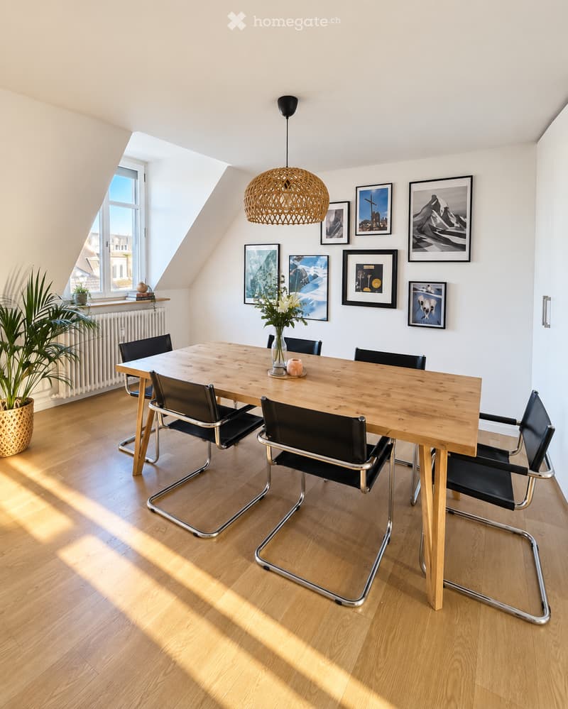 Bright, airy dining area with a wooden dining table surrounded by black chairs, a pendant light, and framed artwork on the walls. A potted plant and radiator are visible near the window.