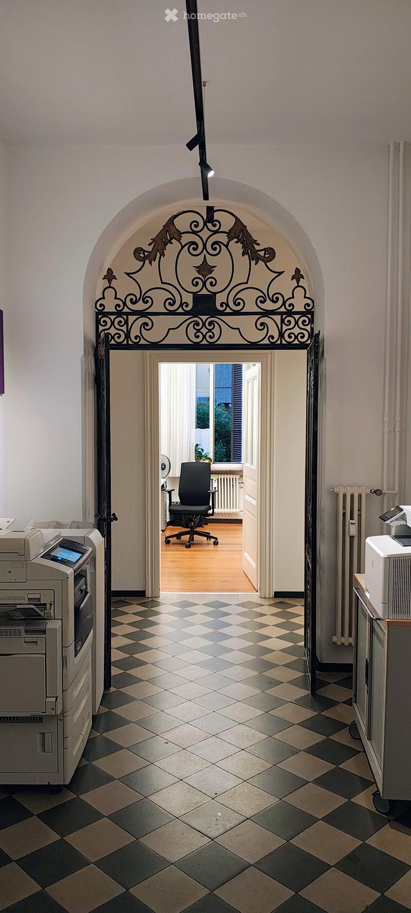 A hallway with checkered black and white tiles, an ornate wrought-iron gate, and a view into an adjacent room with a black office chair and a printer.