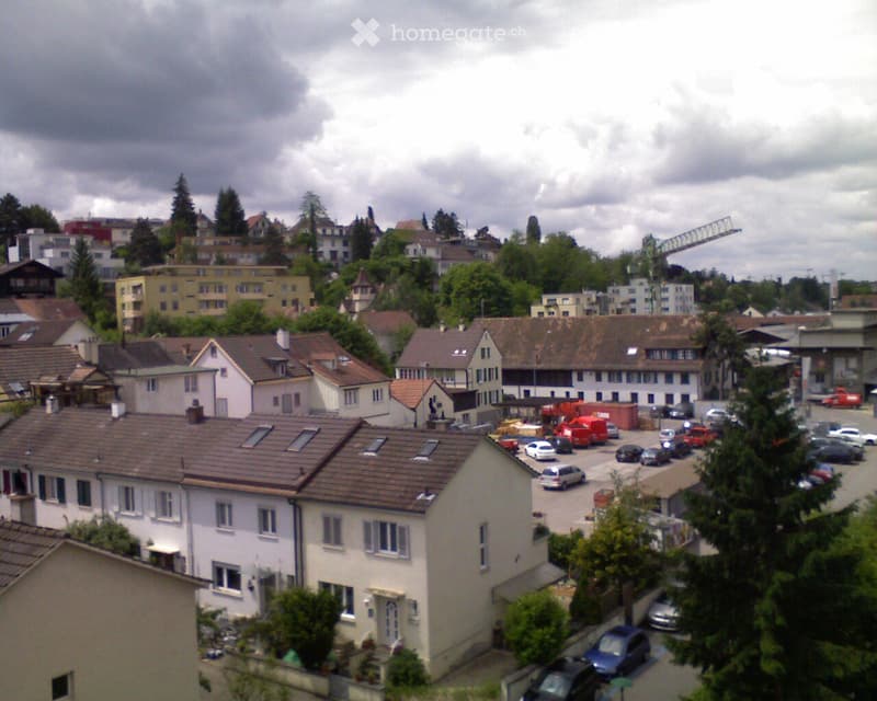 A view of a residential area with multiple houses, parking lot, and trees under a cloudy sky.