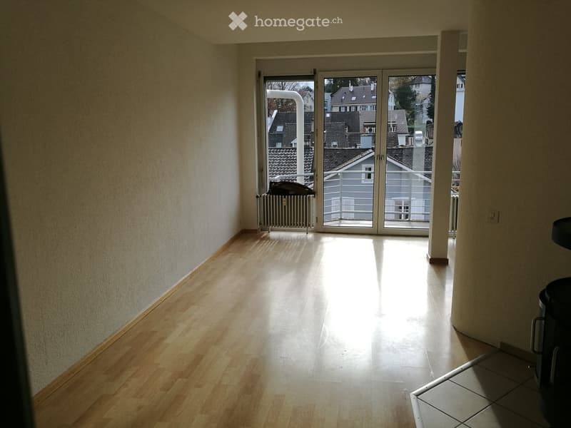 Empty hallway with wooden flooring, white walls, and a large sliding glass door leading to a balcony with a view of rooftops.