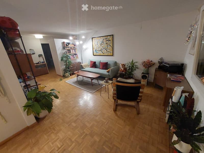 Living room with wooden floor, couch, coffee table, chair, plants, bookshelf, painting, and various decorative items.