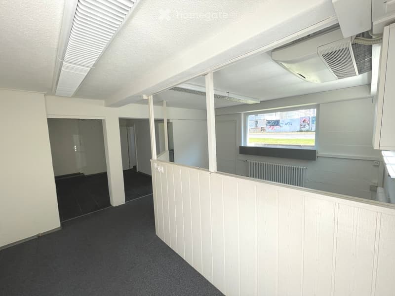 Empty room with white walls, dark carpeted floor, and a large window with a view of the outside. The room features a radiator and a ceiling-mounted air conditioning unit.