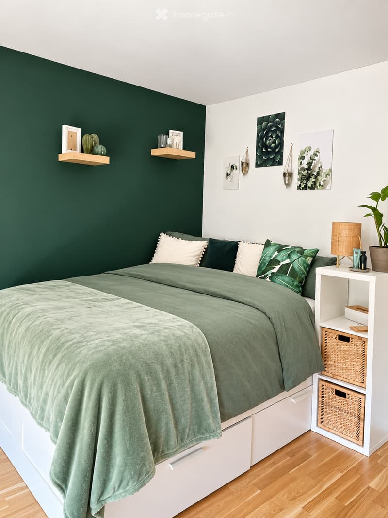 A modern bedroom with a green accent wall, a neatly made bed with green bedding, a white nightstand with a lamp, and decorative shelves with plants and picture frames.