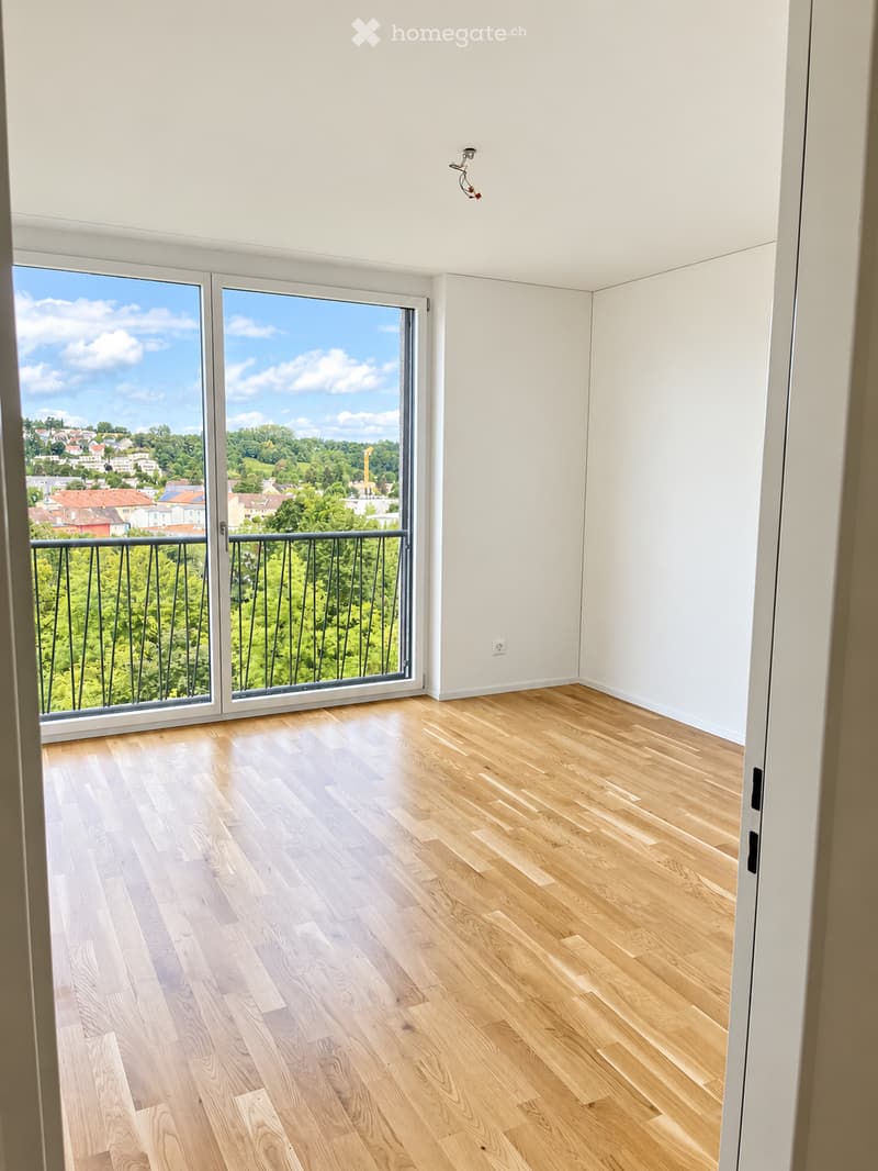 Empty room with light wooden flooring, white walls, and large sliding glass doors leading to a balcony with a view of a cityscape and greenery.