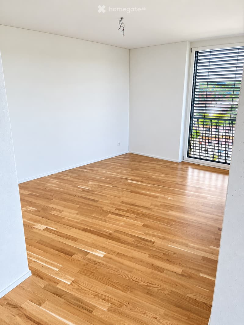 Empty room with light wooden flooring and white walls, featuring a large window with blinds and a view of the outside.
