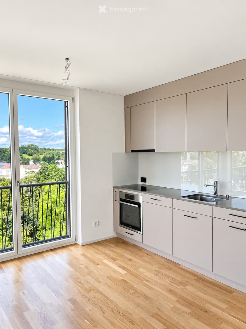 Modern kitchen with light-colored cabinets, stainless steel oven, sink, and faucet, featuring a large window with a view of greenery and a balcony.