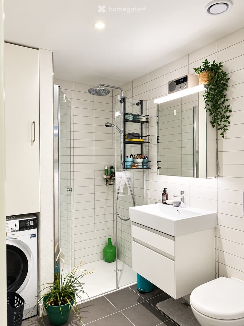 Modern bathroom with white tiles, a glass-enclosed shower, a sink with a mirror, a toilet, a washing machine, and decorative plants.