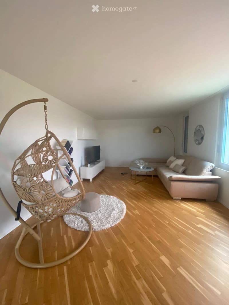 Modern living room with light wooden flooring, white walls, a beige sofa, a wicker hanging chair, a coffee table, and a TV stand.