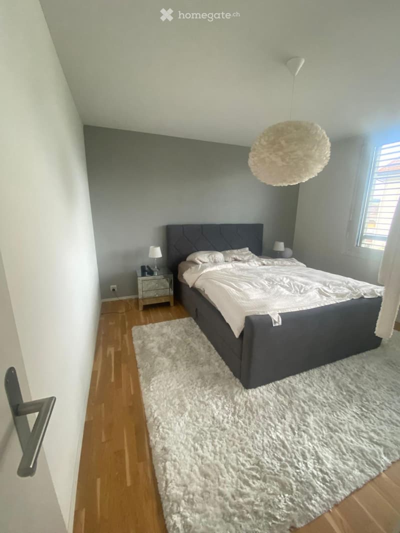 Modern bedroom with a dark grey upholstered bed, white bedding, a fluffy white rug, wooden nightstand, and pendant light.