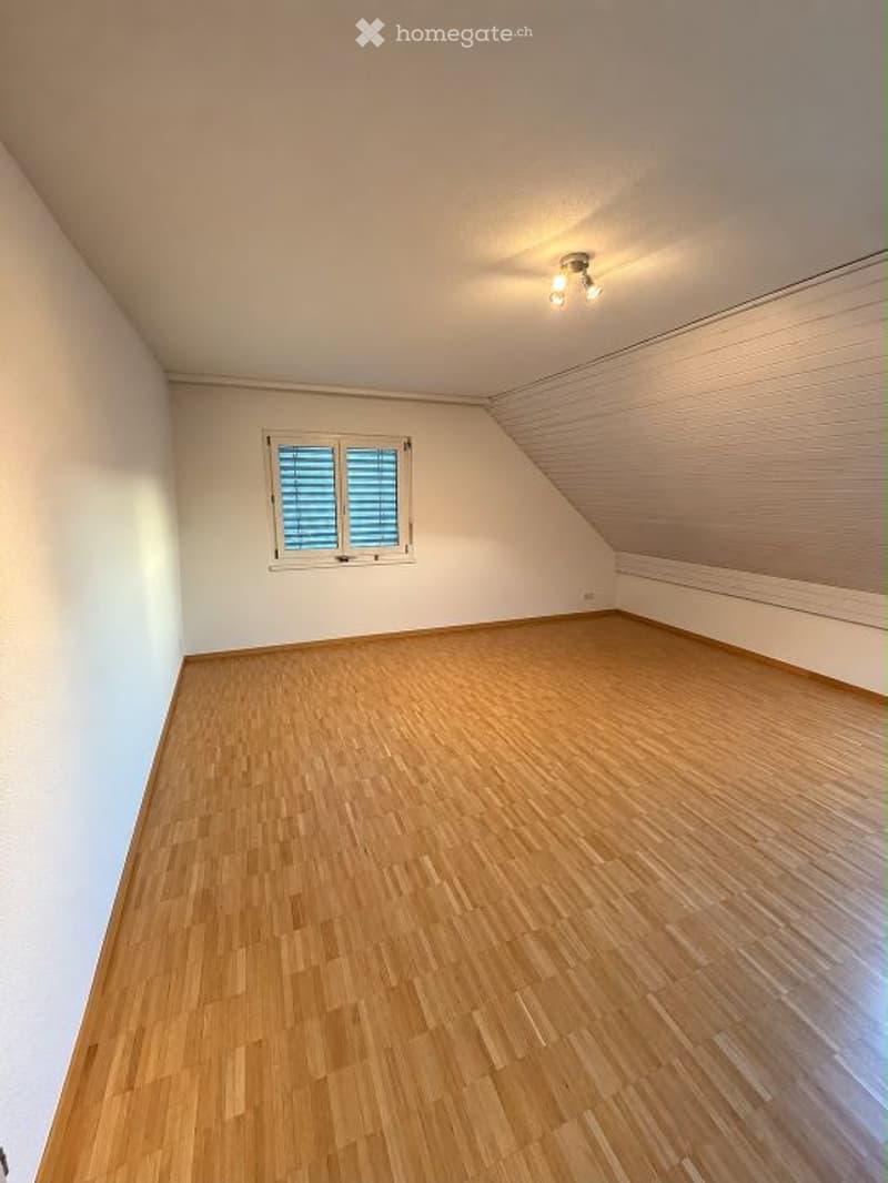 Completely empty attic room with wooden flooring, white walls, and a single window with blinds. The ceiling has a sloped design with integrated lighting.