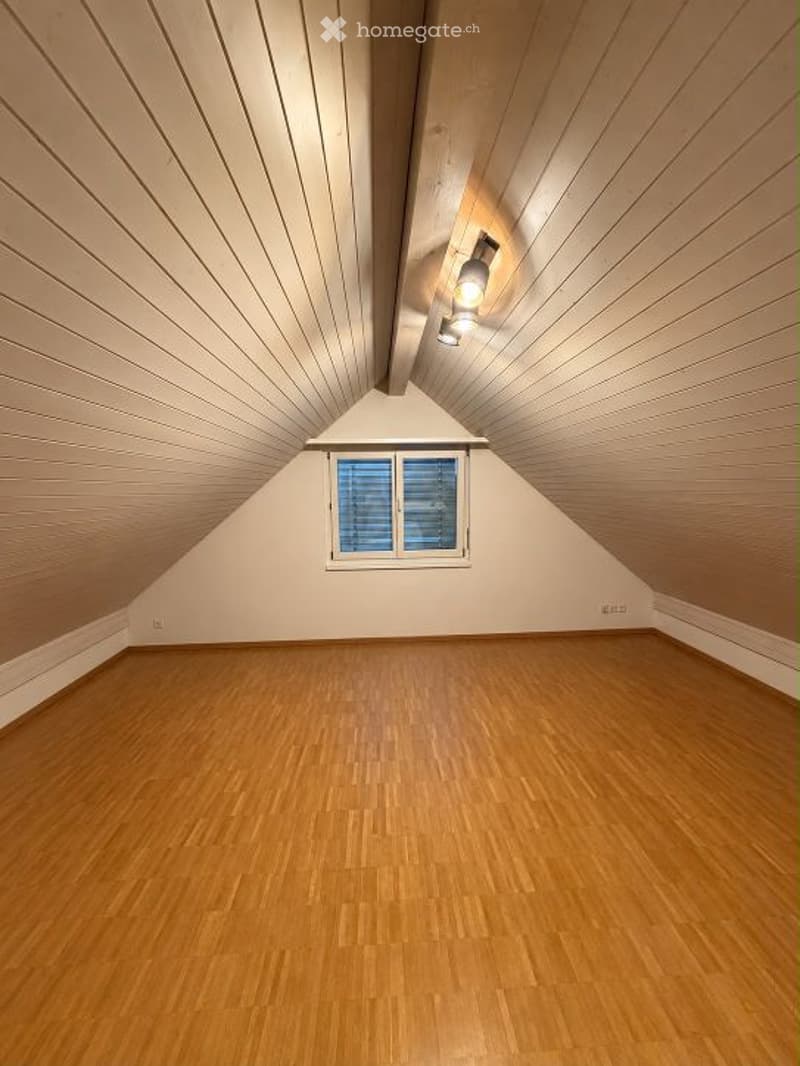 Empty attic room with wooden floors, slanted wooden ceiling, and a single window with blinds.