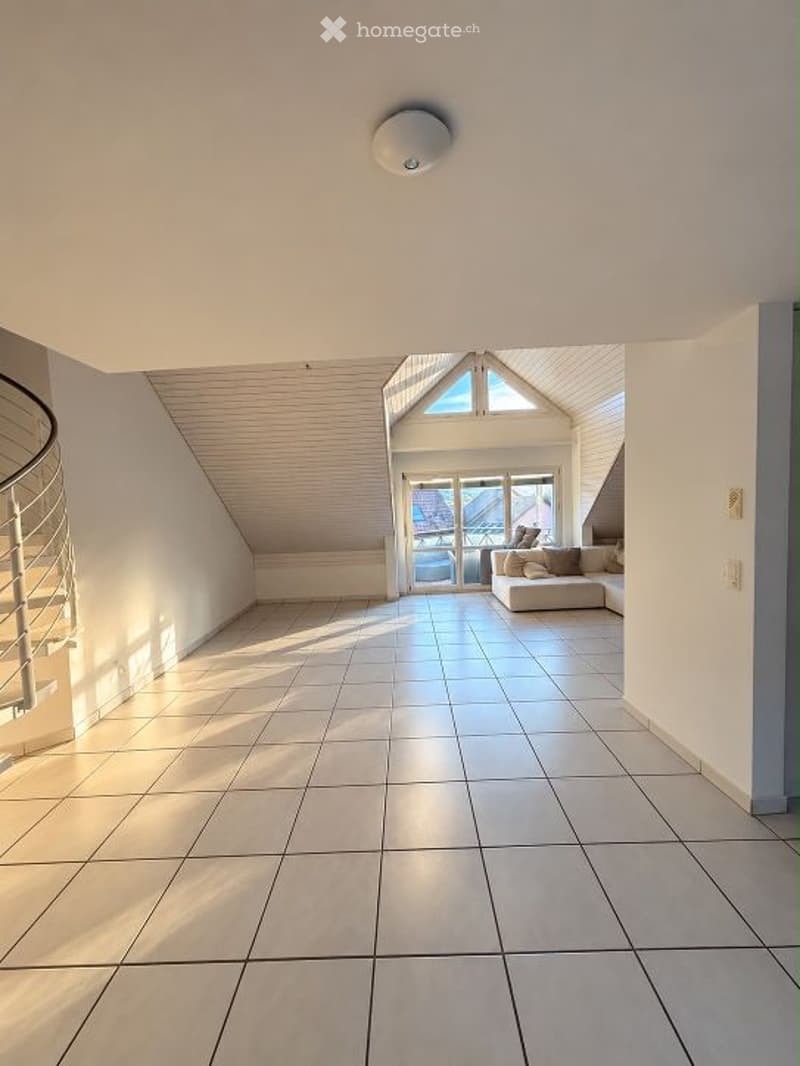 Bright, open space with white tiled flooring, a modern white sofa with pillows, and large windows offering a view of the outside. The room features a sleek, curved staircase on the left and a skylight above.