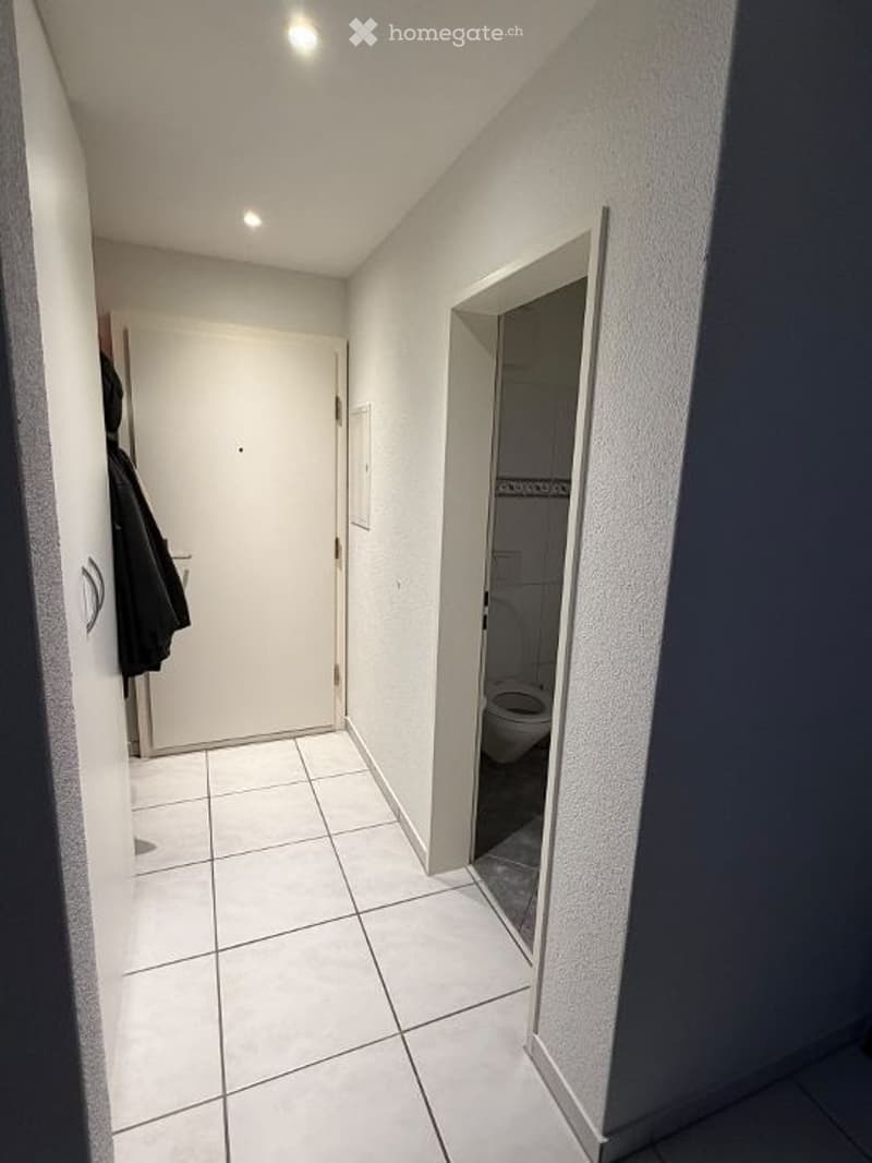 A narrow, well-lit hallway with white tiled flooring and light grey walls. A closed white door is on the left, and an open door on the right leads to a small bathroom with a visible toilet.